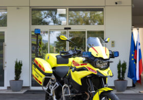 Novo motorno vozilo nujne medicinske pomoči ZD Bled, parkirano pred vhodom zdravstvenega doma z izpisom "POMOČ ZD BLED" in državnimi zastavami v ozadju.